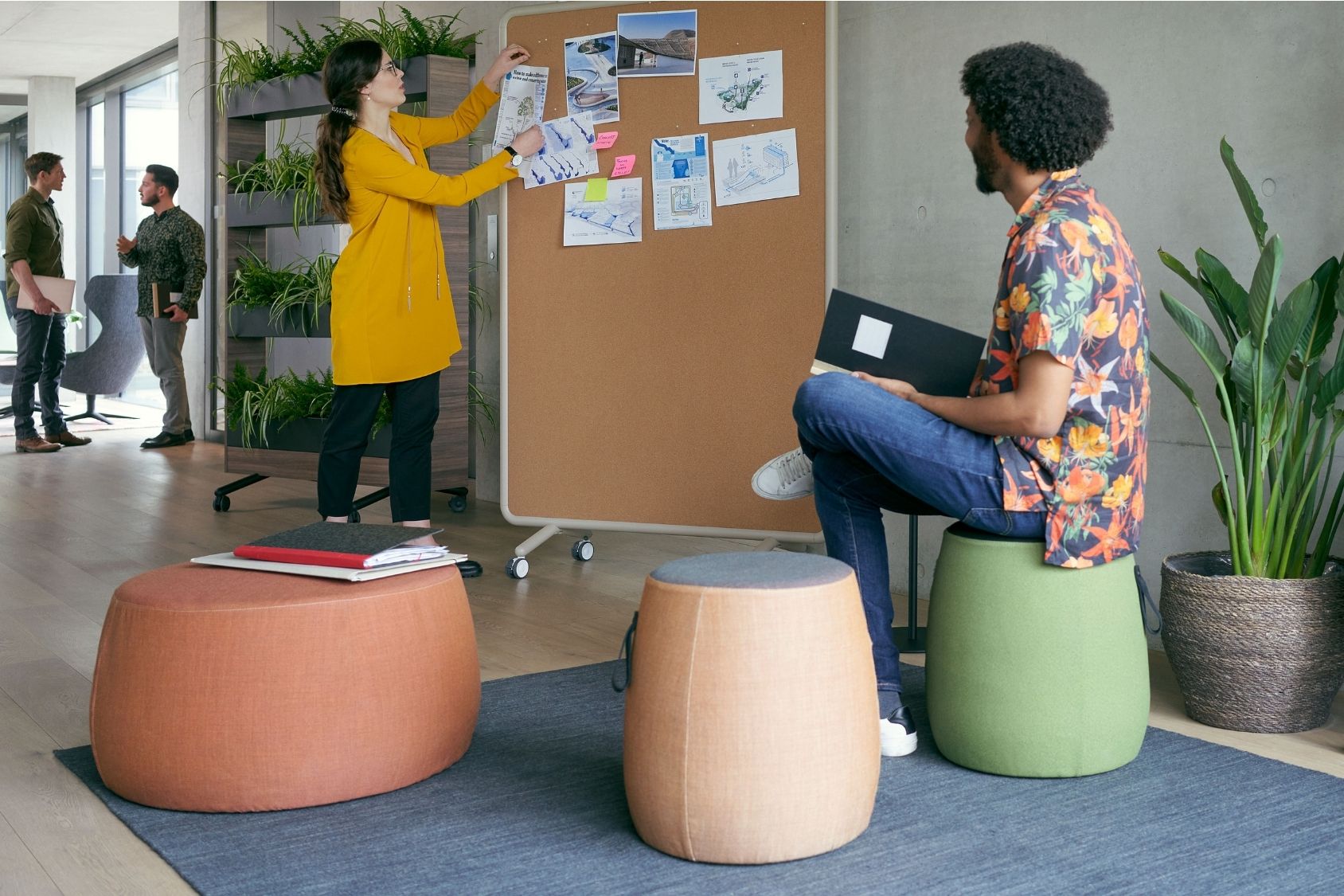 Drei se:dot Hocker in Orange, Beige und Grün mit rundem, gepolstertem Design in einem modernen Büro. Frau in gelbem Mantel pinnt Notizen an eine Pinnwand. Mann mit floralem Hemd sitzt auf grünem Hocker und hält schwarzen Ordner.