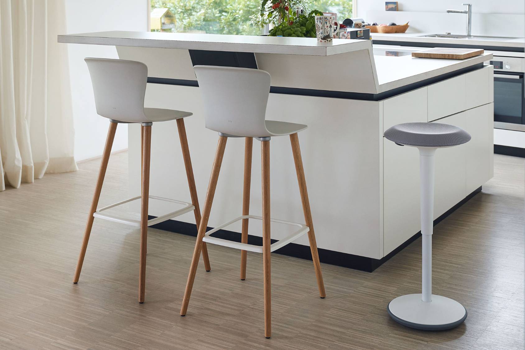 Zwei se:spot stool Barhocker mit weißen Sitzflächen und Holzbeinen an einer Kücheninsel, daneben ein se:fit in grauer Ausführung.
