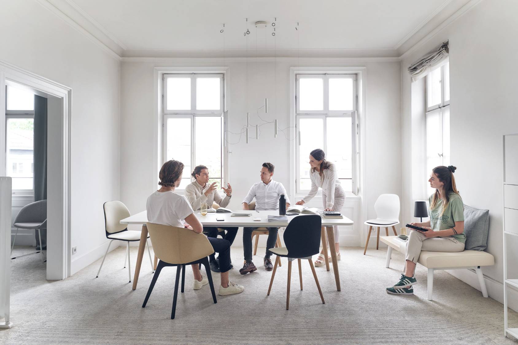 Fünf Personen sitzen um einen weißen Tisch mit verschiedenen Stühlen, darunter der se:spot Stuhl mit ergonomischem, modernem Design und Holzbeinen, in einem hellen, minimalistischen Raum mit großen Fenstern.