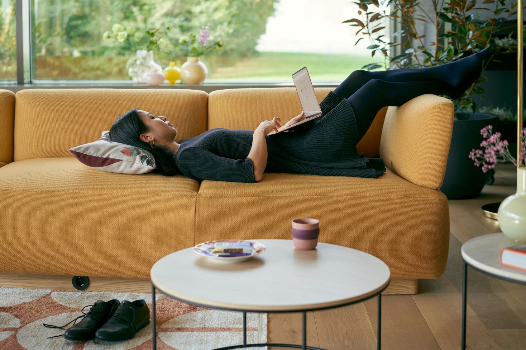 se:living Sofa in senfgelb, modernes Design mit klaren Linien und abgerundeten Ecken, Zweisitzer mit Rollen. Person liegt entspannt mit Laptop darauf.