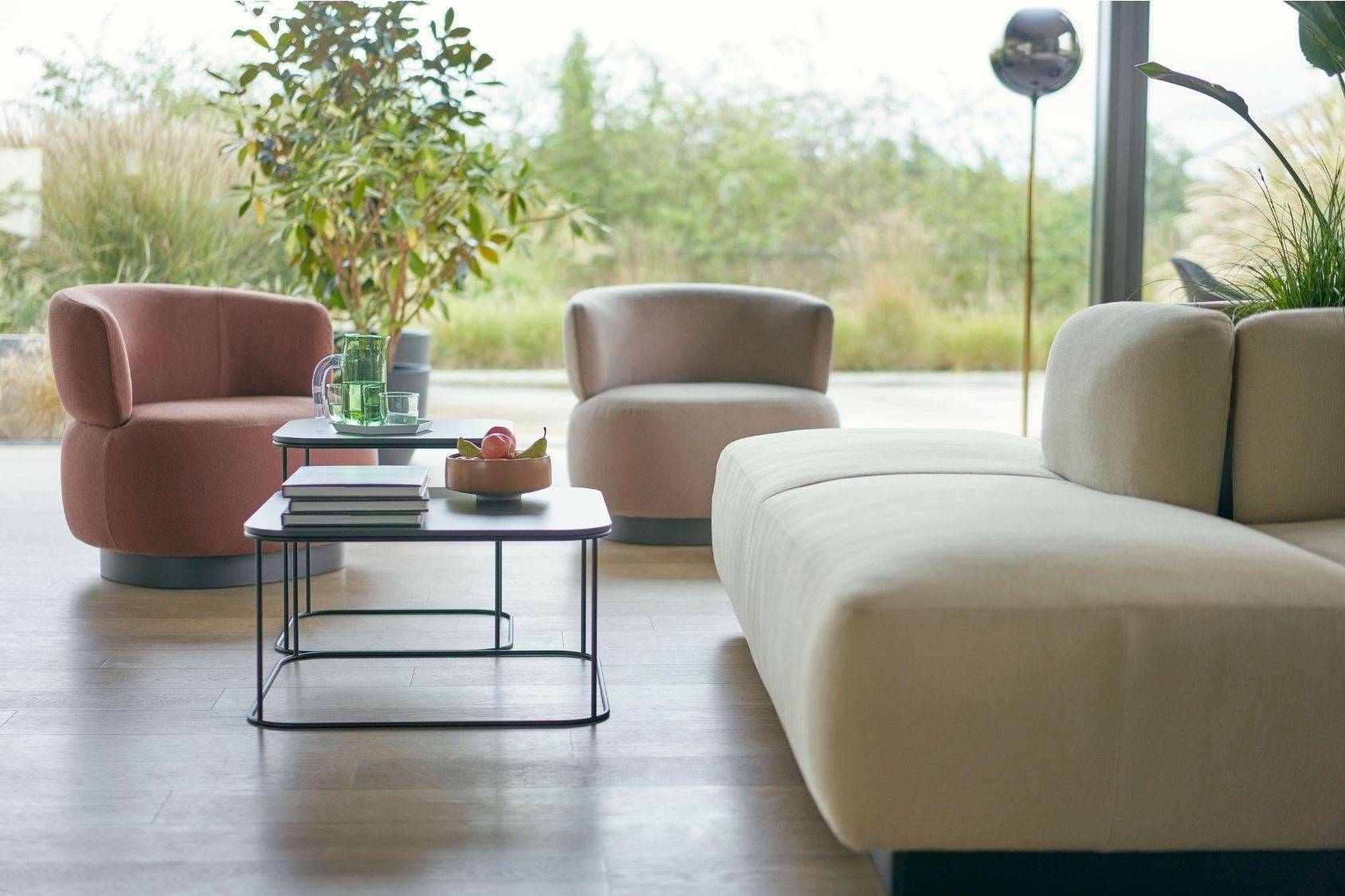 se:living Sessel in Rot und Beige neben einem beigen Sofa und zwei schwarzen Couchtischen mit Büchern und Dekoration in einem hellen Wohnzimmer mit Blick ins Grüne