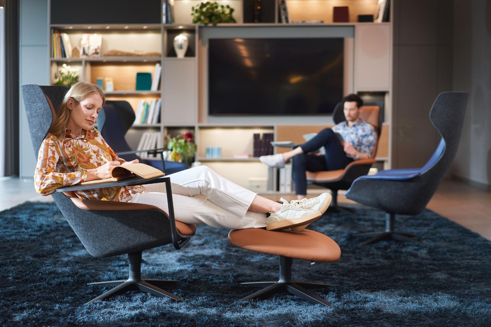 se:lounge Ottoman Fußhocker in braun vor einem grauen Sessel, eine Person sitzt entspannt mit ausgestreckten Beinen darauf in einem modernen Wohnzimmer