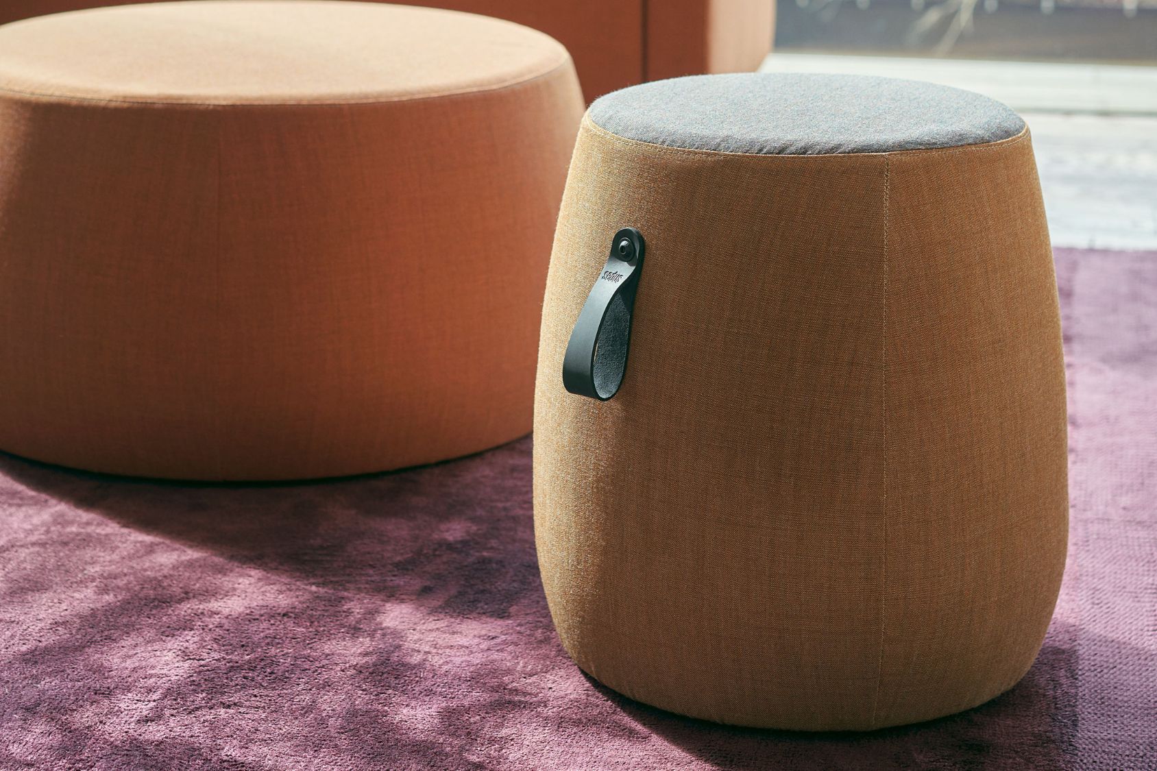 se:dot Hocker mit zylindrischem, abgerundetem Design und Stoffbezug, braun mit schwarzem Griff, neben rundem orangefarbenem Hocker auf lila Teppich