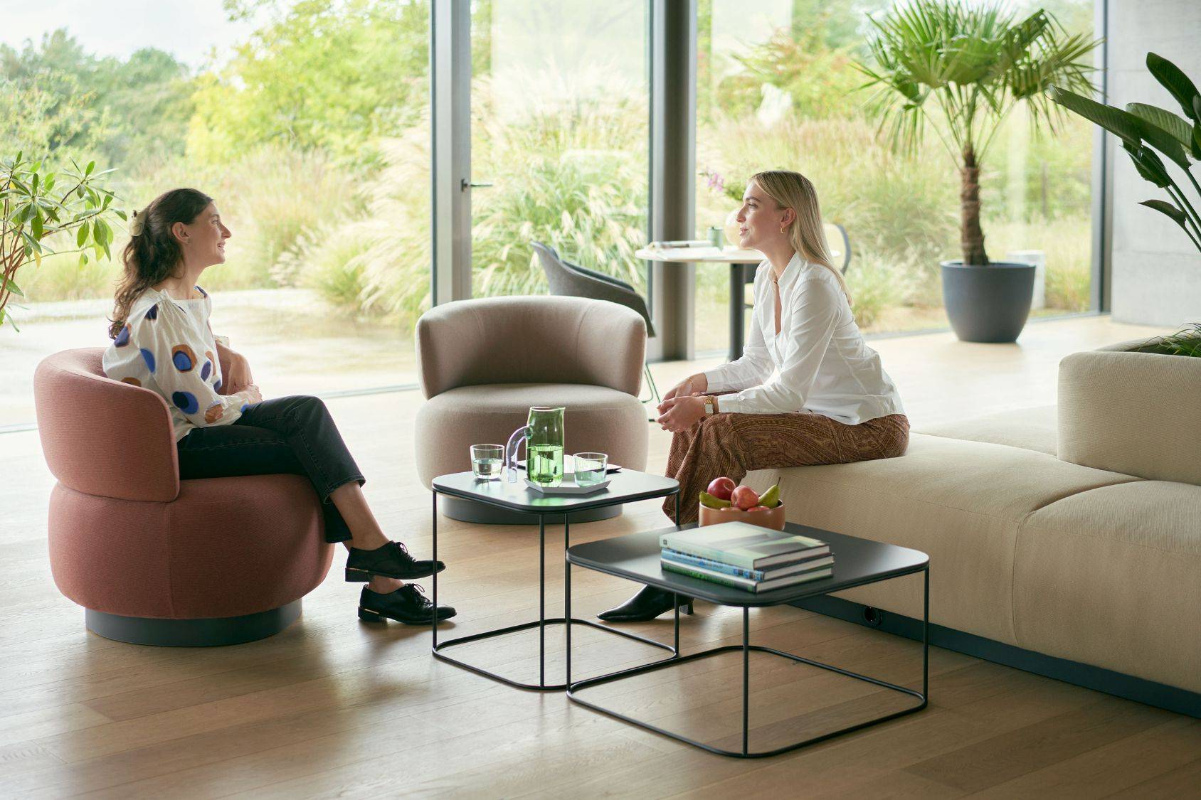Zwei Frauen sitzen im Wohnzimmer, eine auf einem se:living Sessel in Rosa, die andere auf einem hellen Sofa, vor großen Fenstern mit Blick ins Grüne