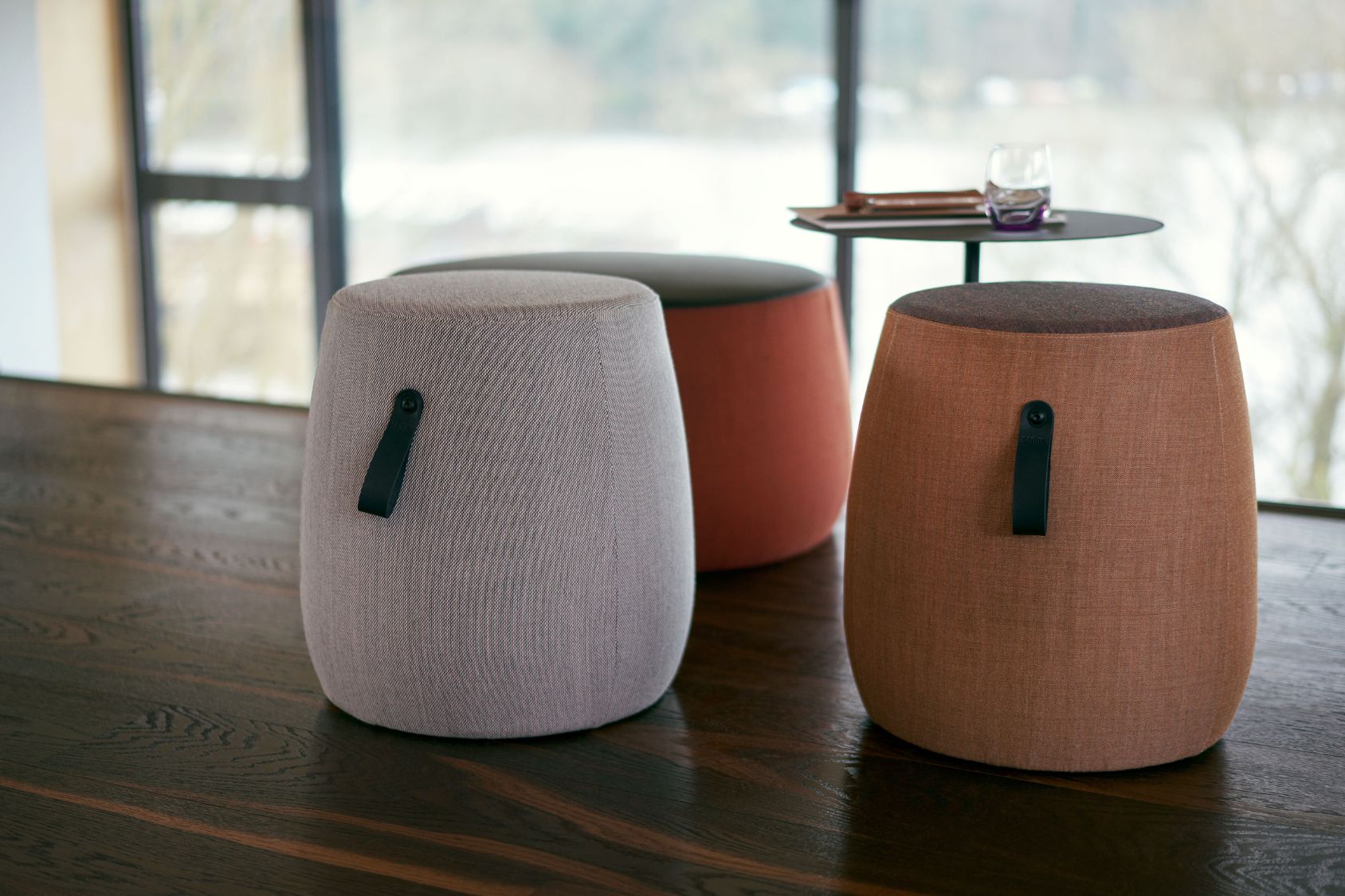 Drei se:dot Hocker mit zylindrischer Form, gepolstert und mit Stoffbezug in Grau, Orange und Braun, jeweils mit schwarzem Tragegriff