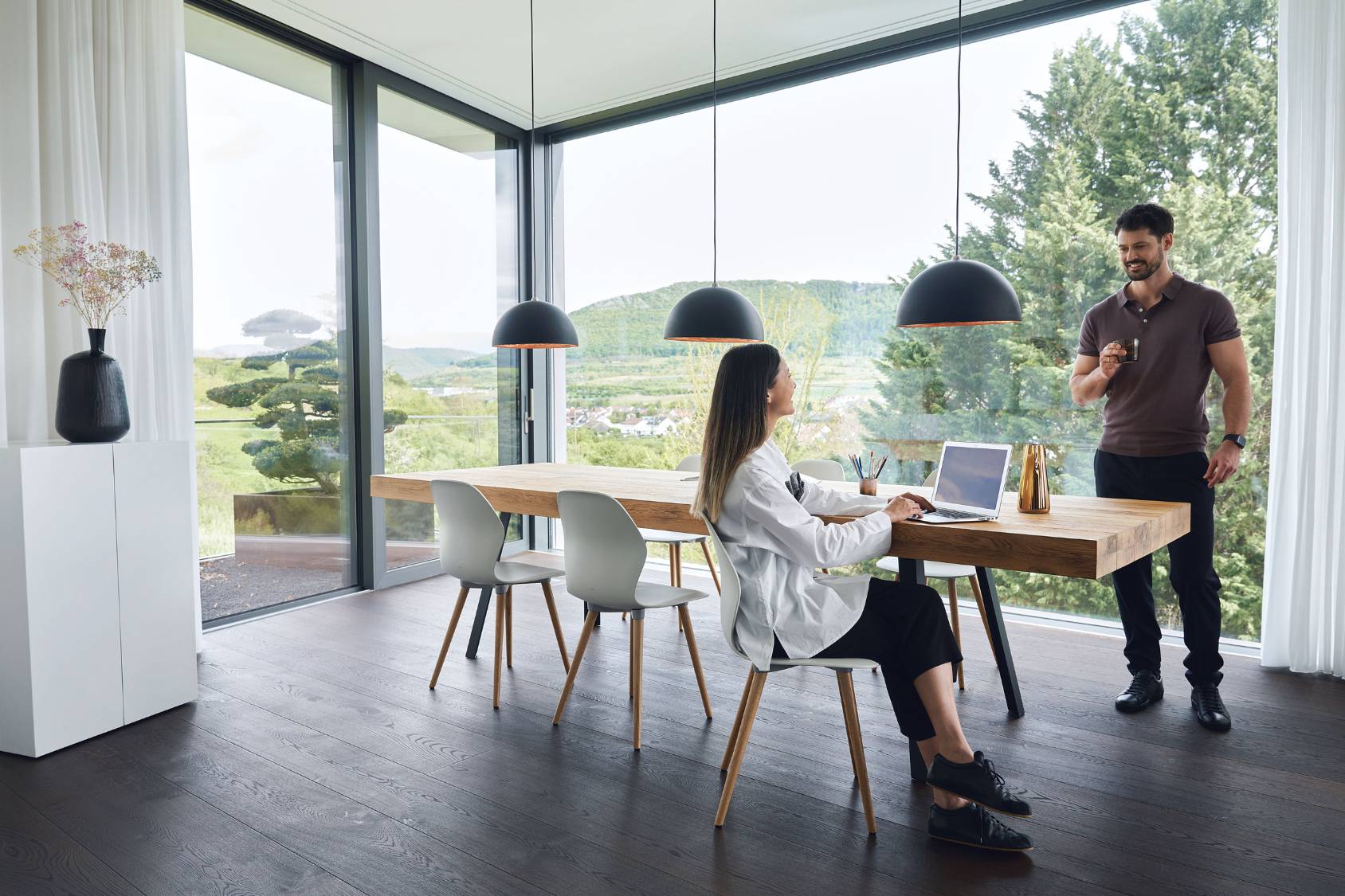 Moderner Essbereich mit großem Holztisch und drei schwarzen se:spot Pendelleuchten mit rundem Design, zwei Personen im Gespräch, eine sitzt mit Laptop am Tisch
