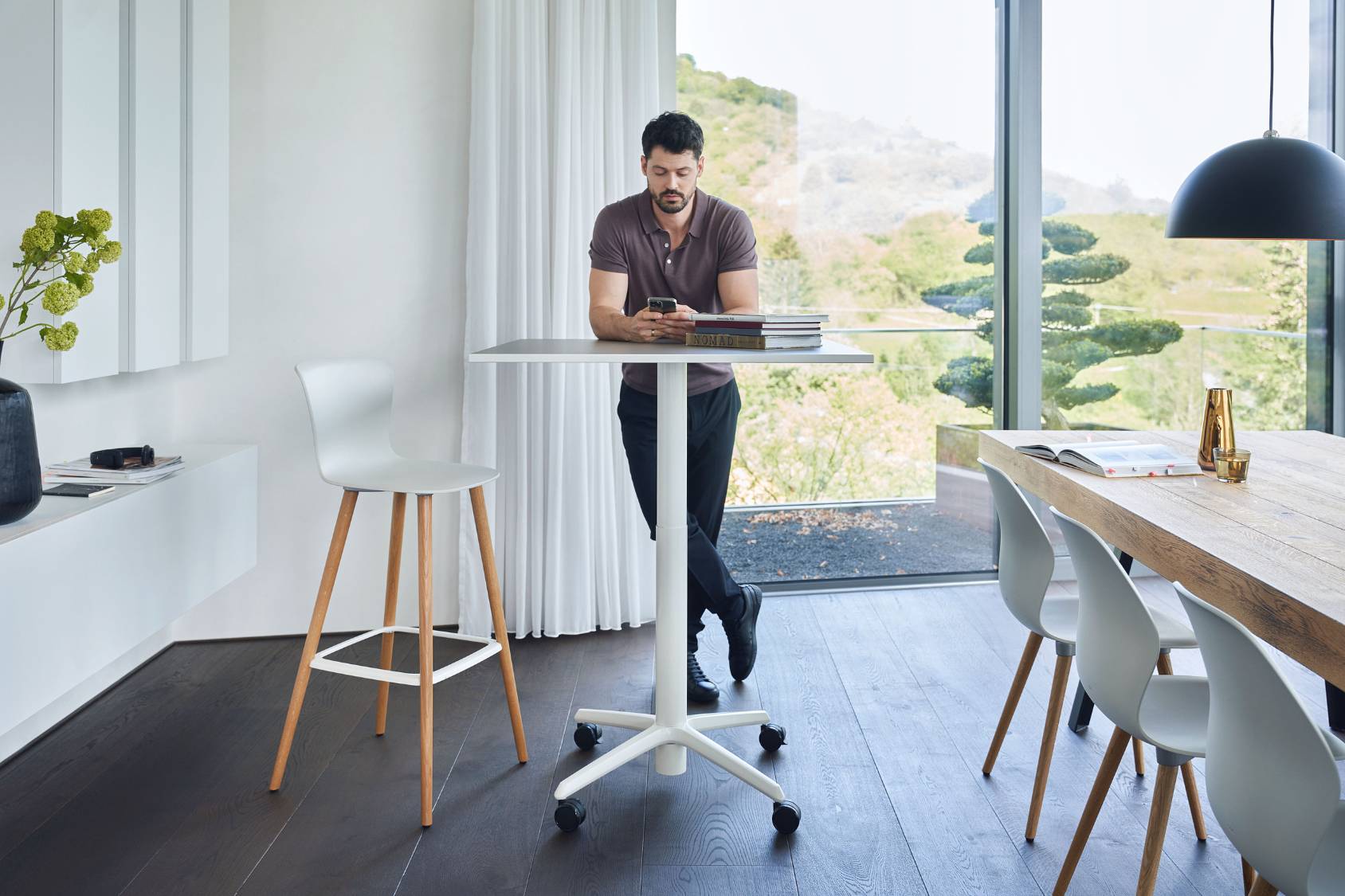 se:spot stool Barhocker mit weißem Sitz und Holzbeinen, modernes minimalistisches Design, steht neben weißem Stehtisch mit Rollen in hellem Raum mit großen Fenstern