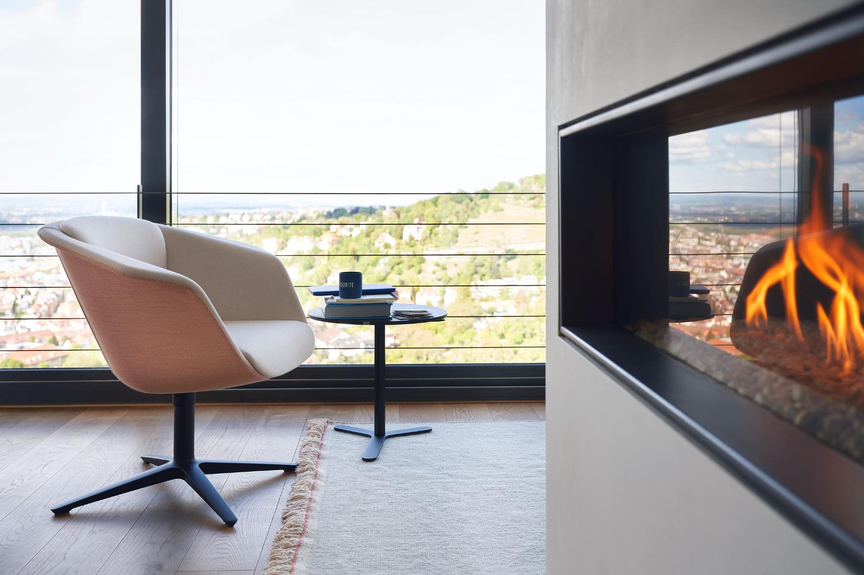 Der sweetspot in Beige mit schwarzem Fußgestell steht neben einem kleinen schwarzen Beistelltisch vor einer Glasfront mit Blick auf eine Stadt und einen Kamin mit Flammen
