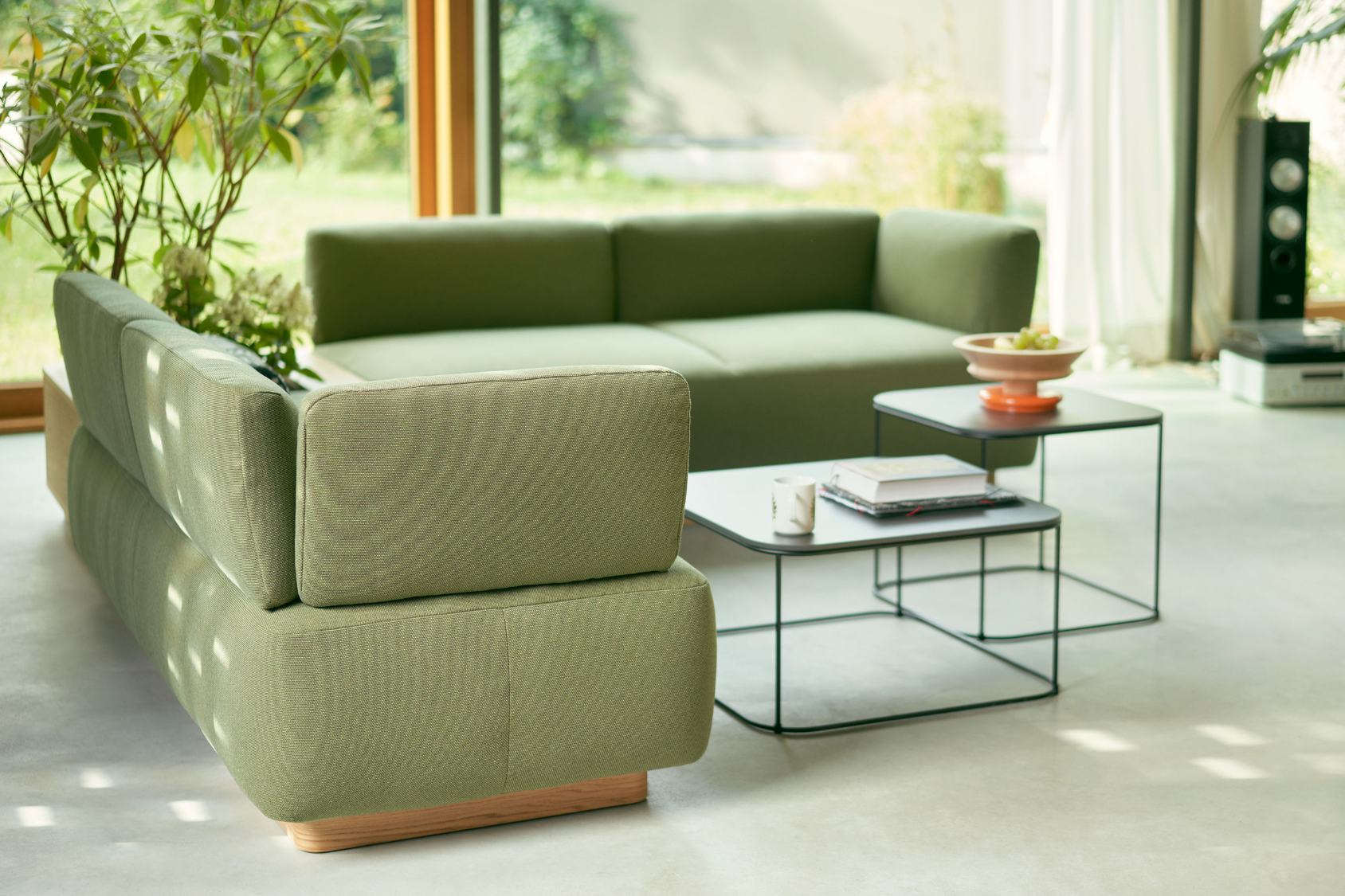 se:living Sofa, zweisitzig, modernes Design mit klaren Linien und grünem Stoffbezug, rechteckige Form, Holzsockel, im hellen Wohnzimmer mit Pflanzen und zwei schwarzen Metall-Couchtischen