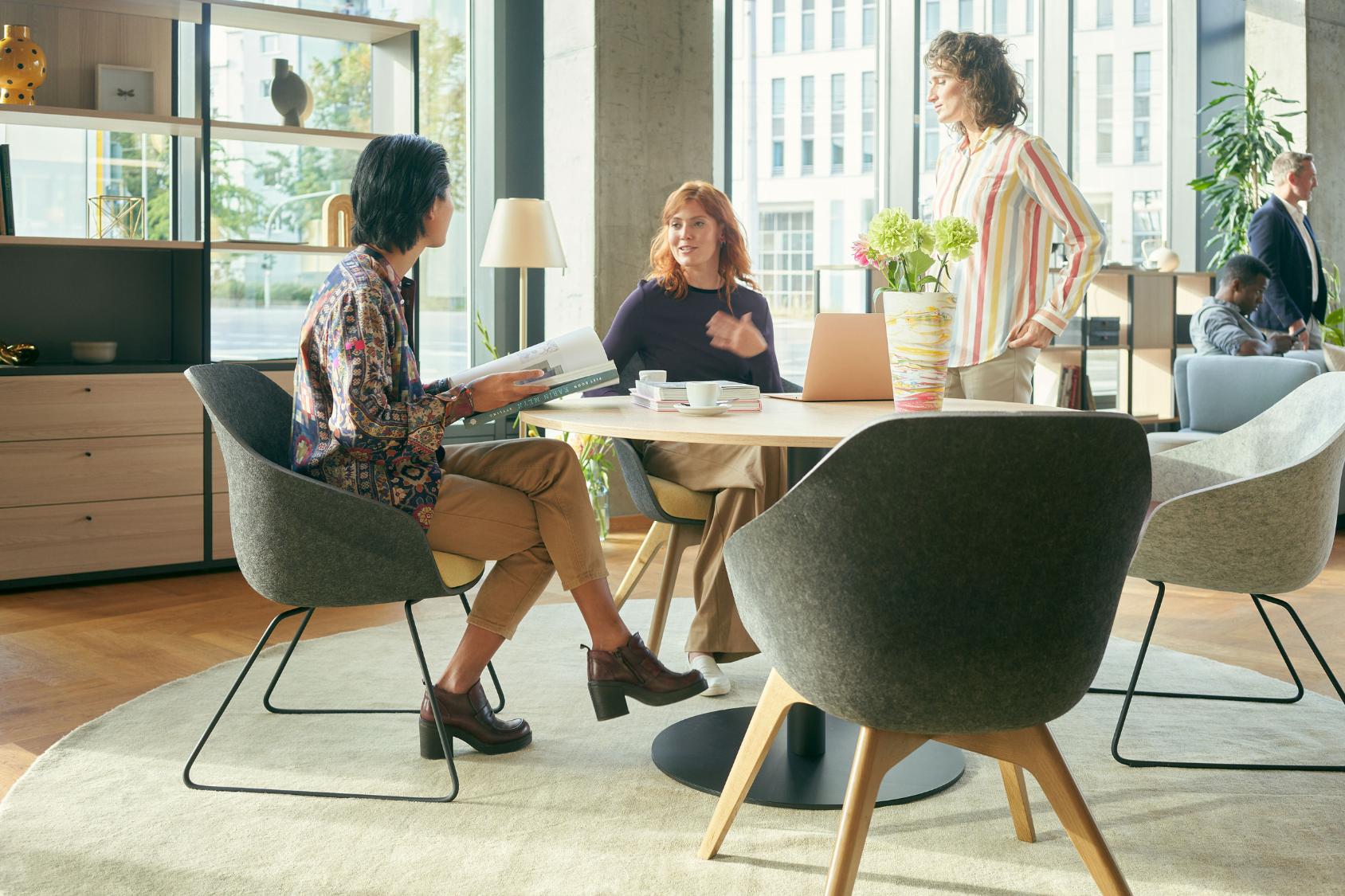 Drei Personen sitzen und stehen um einen runden Tisch in einem modernen Büro, alle nutzen den se:lounge light Bürostuhl mit gepolstertem Sitz und schlanken Beinen.