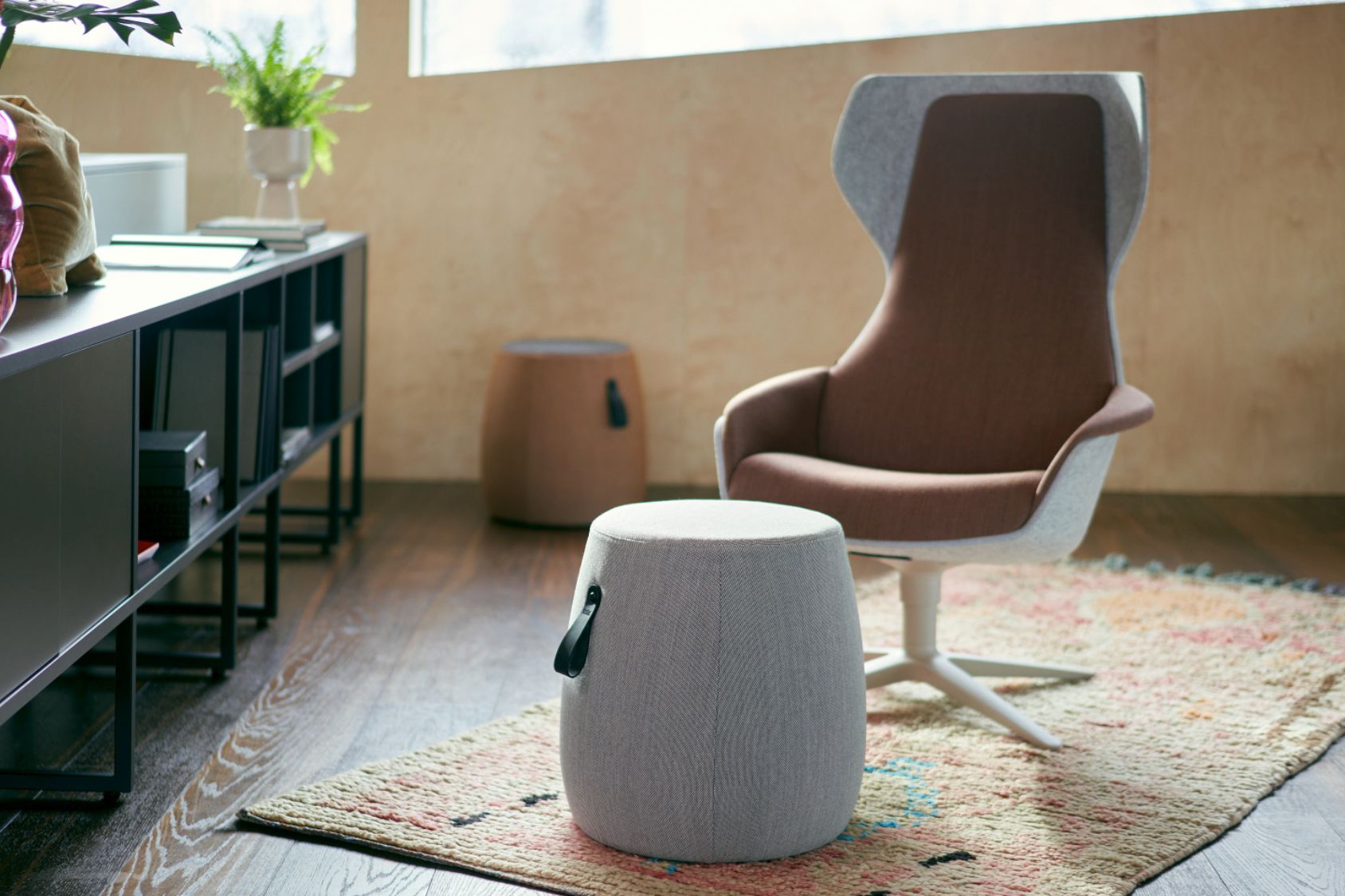 se:lounge Sessel mit hohem Rücken und ergonomischem Design in Braun, davor grauer runder Hocker mit Tragegriff, beides auf gemustertem Teppich in modernem Wohnzimmer